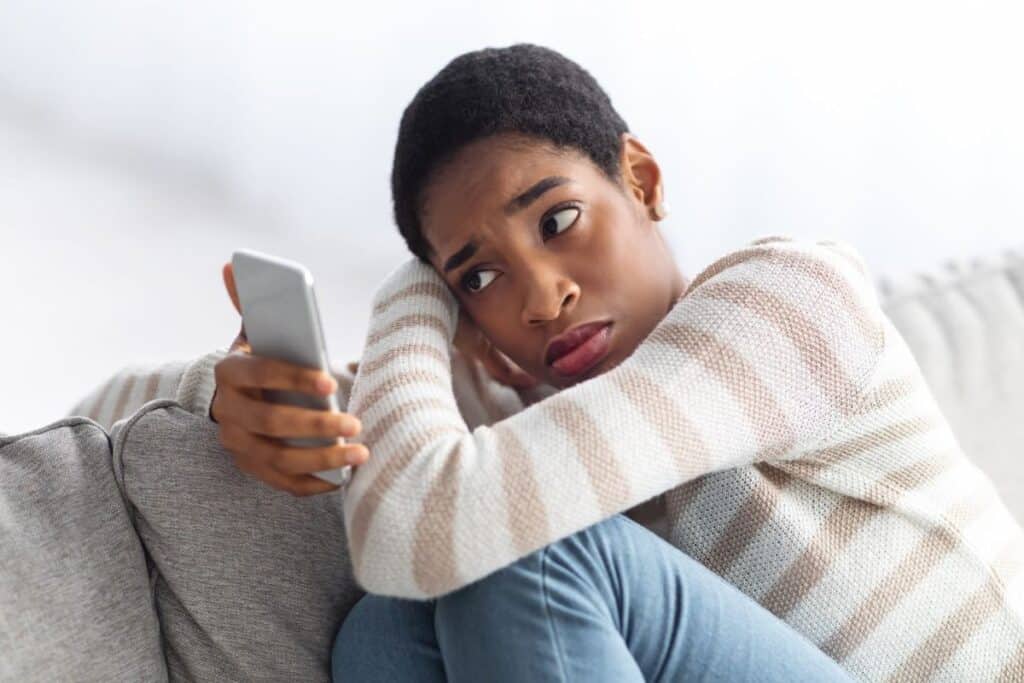 A young African-American woman worriedly looks at her smartphone while hugging her knees.